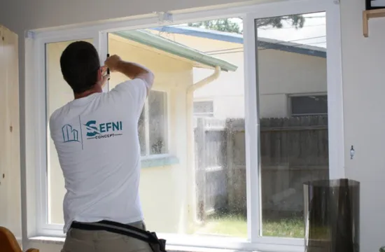 Personne installant un film pour vitres sur une fenêtre à triple vitrage ; vue extérieure ensoleillée.